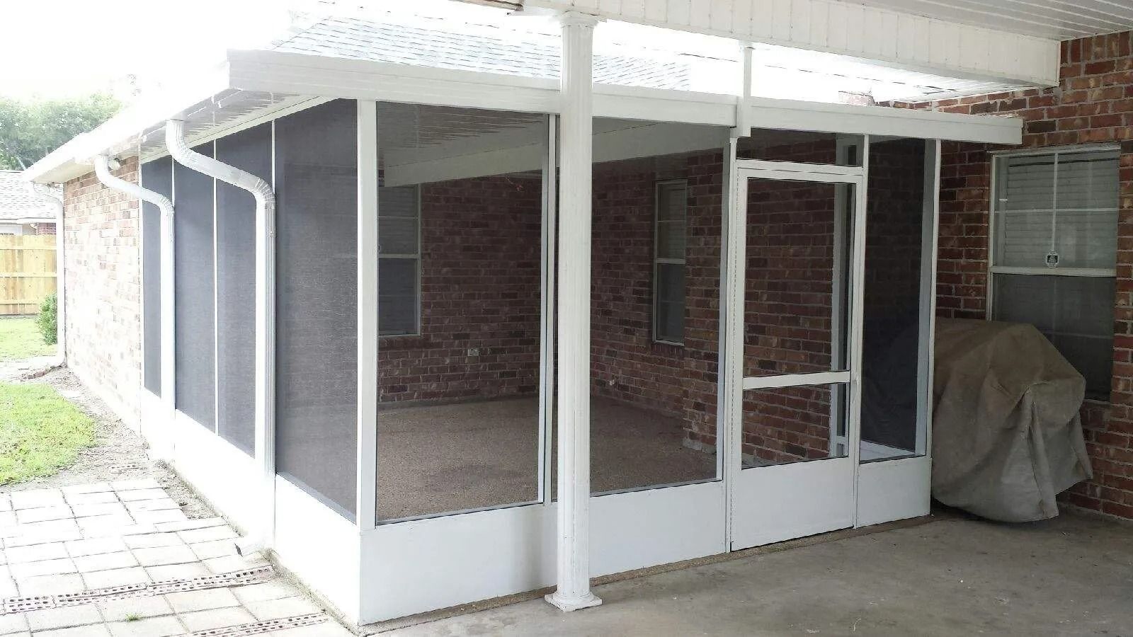 Screened-in porch attached to a brick house. White frame with dark screens, clear roof, and a door.
