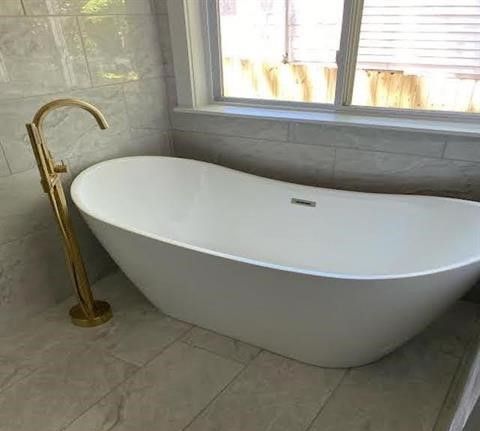 A white bathtub with a gold faucet in a bathroom next to a window.
