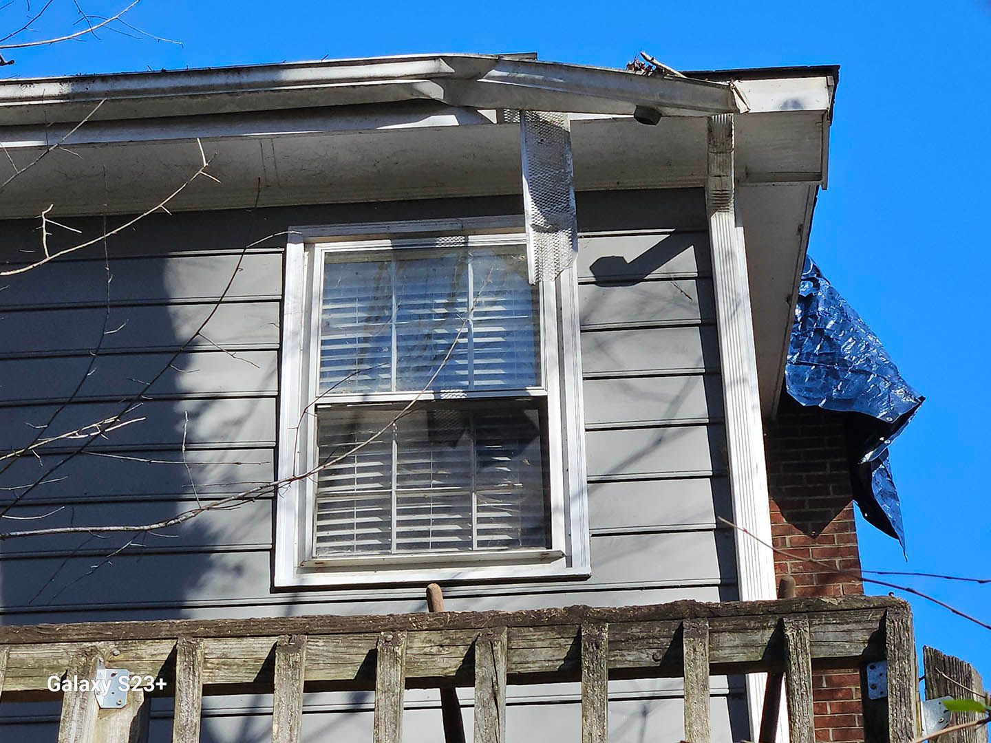 A gray house with a window and a blue tarp on the roof