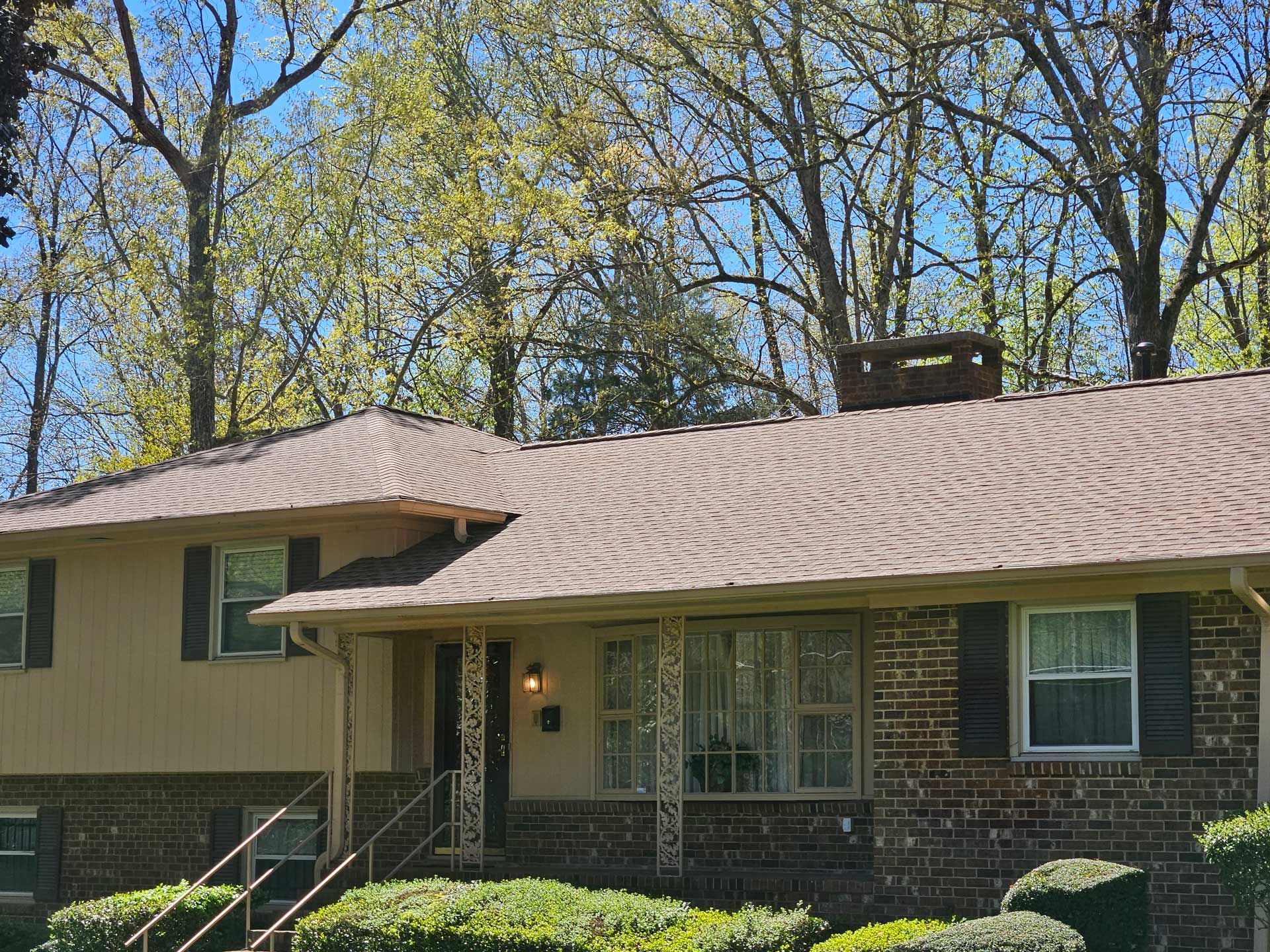 A brick house with a brown roof and black shutters is surrounded by trees.