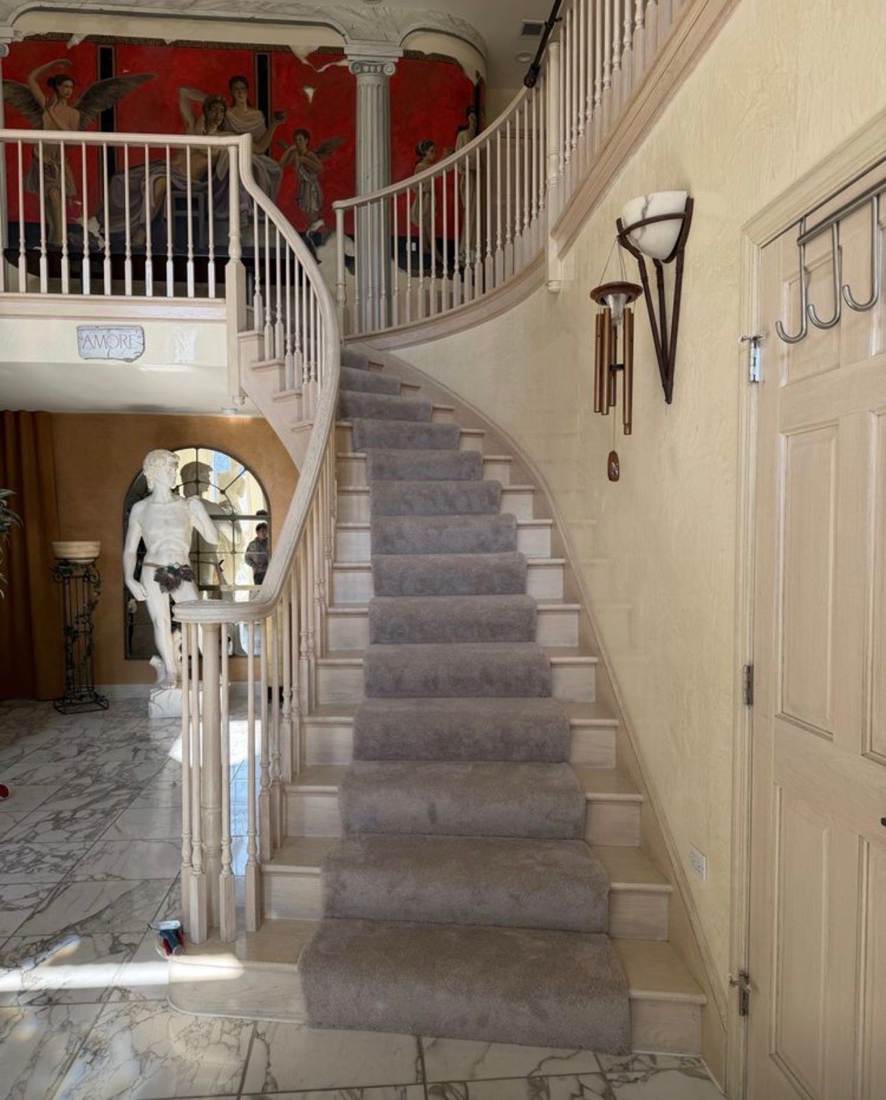 Curved gray-carpeted staircase in a grand foyer with red wall mural and beige doors.