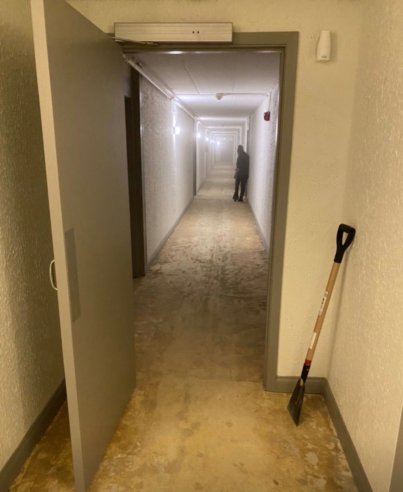 Long apartment hallway under renovation, with a person in the distance and a shovel leaning by the door