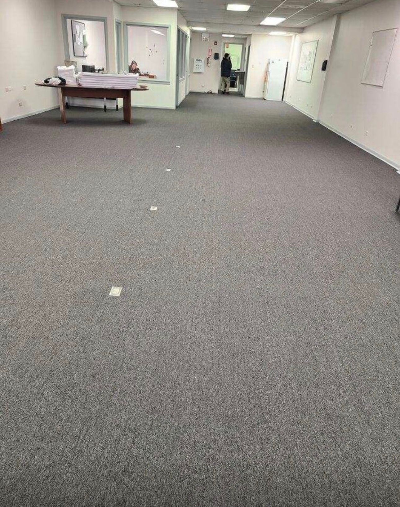 Empty carpeted office room with white walls, fluorescent lights, and a table near the left wall.