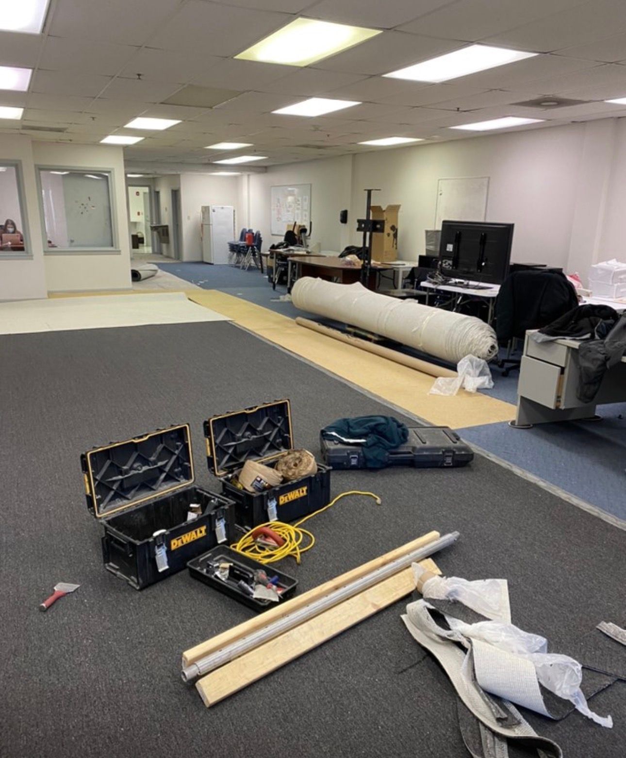 Office with scattered tools and supplies on carpet, a rolled material on the floor, and desks along the wall.