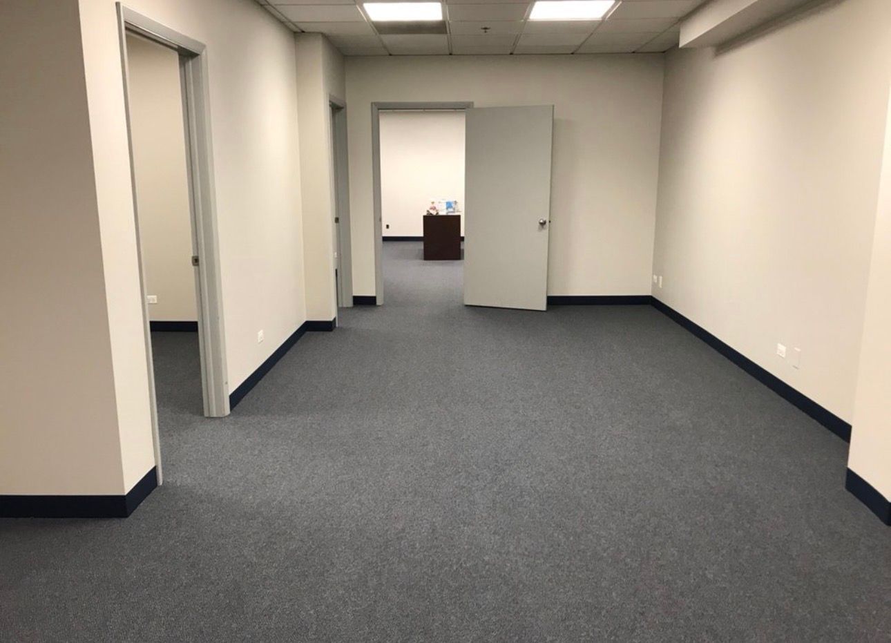 Empty office hallway with blue carpet, beige walls, and fluorescent ceiling lights leading to a doorway.