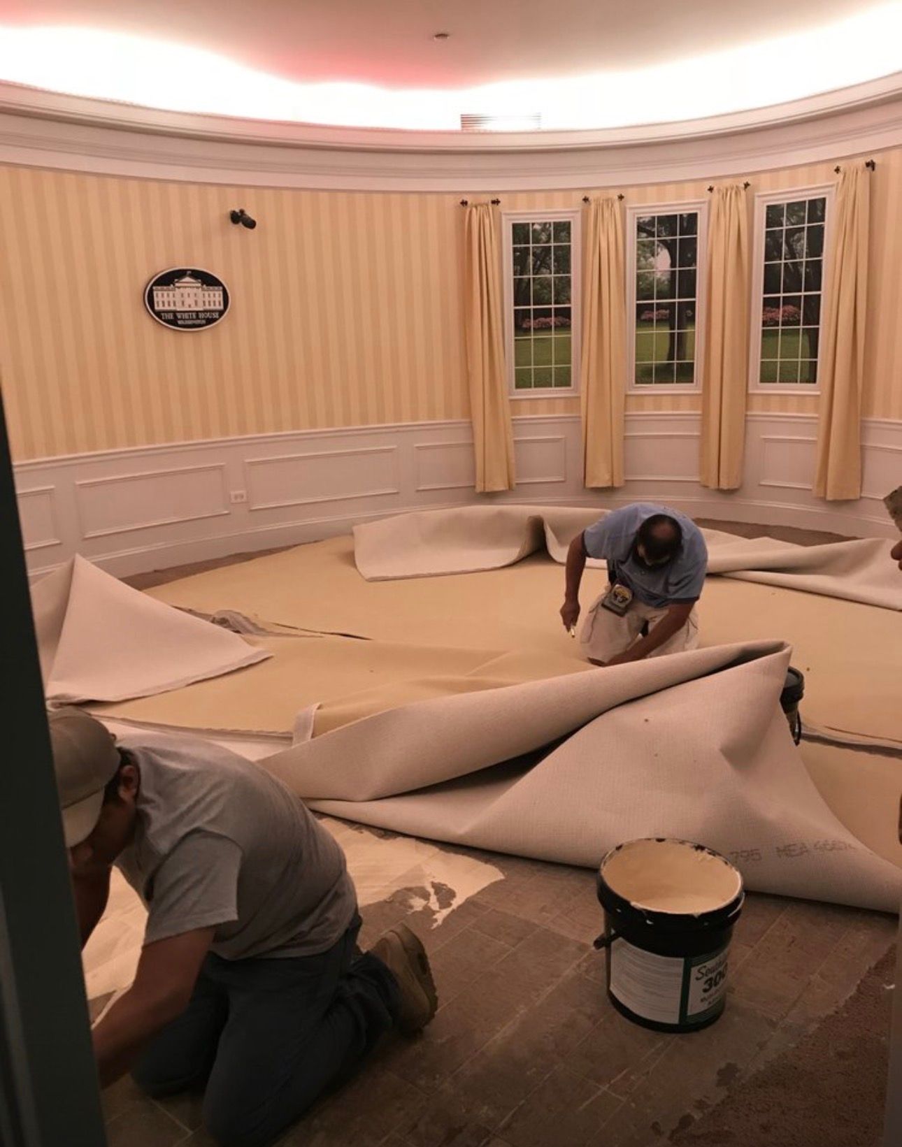 Workers roll up carpet in a room with beige walls and curtained windows.