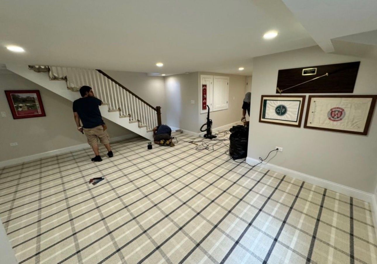 Bright basement room with white grid tile floor, staircase, framed art, and two people working inside.