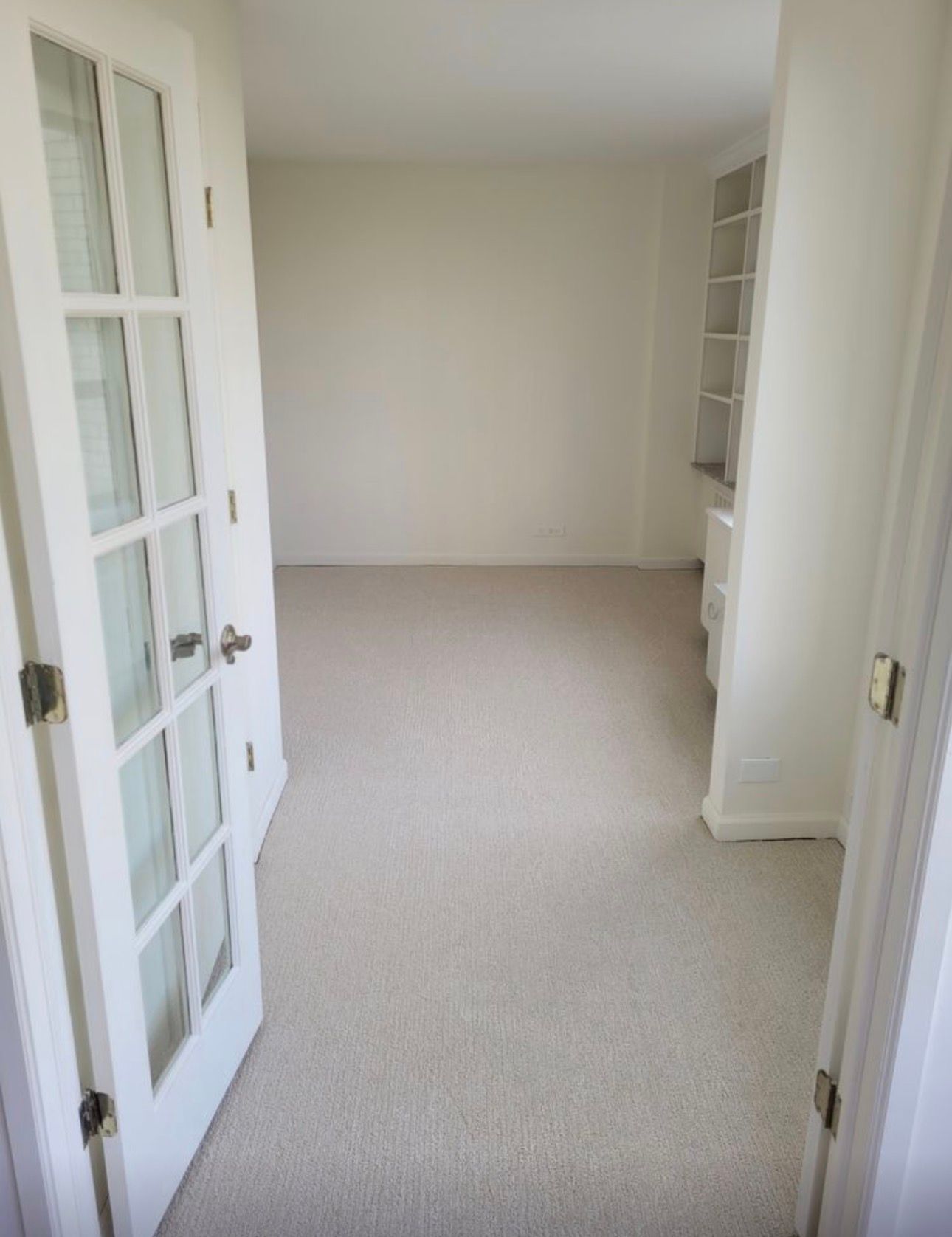 Empty beige room with carpet, white French doors, and built-in shelving visible through the doorway.