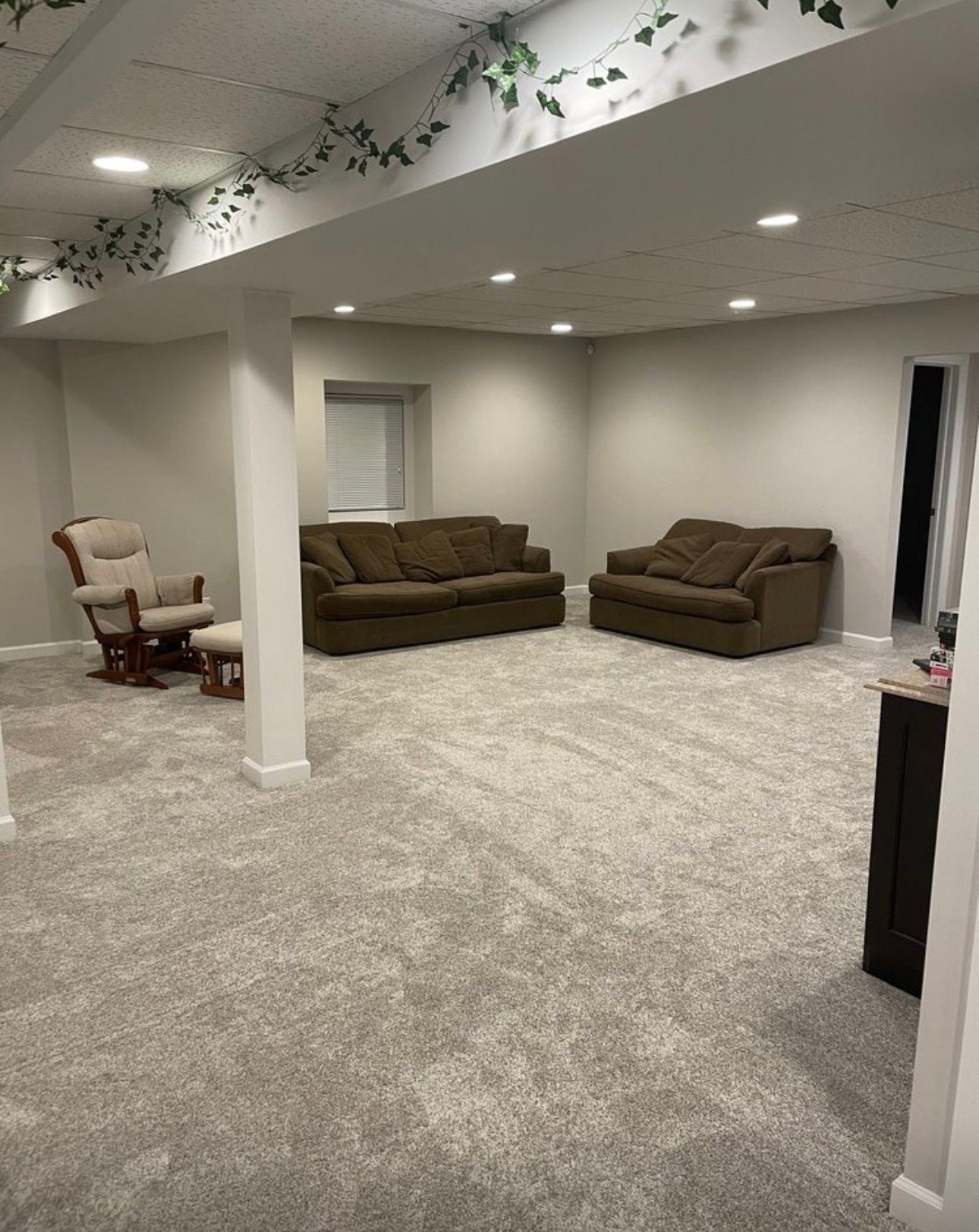 Finished basement family room with carpet, brown sofas, a rocking chair, and recessed lighting.