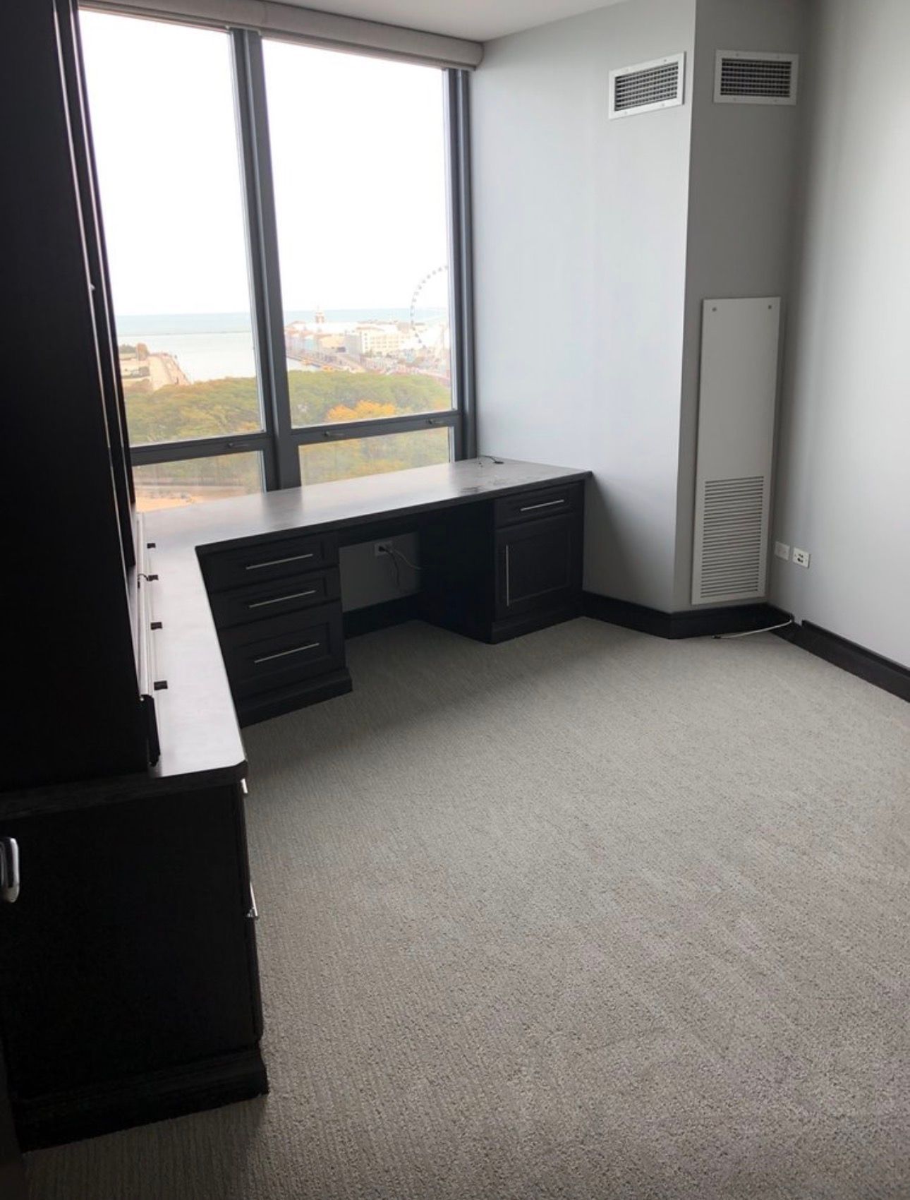Empty office with gray carpet, black desk, and large windows overlooking a city view.
