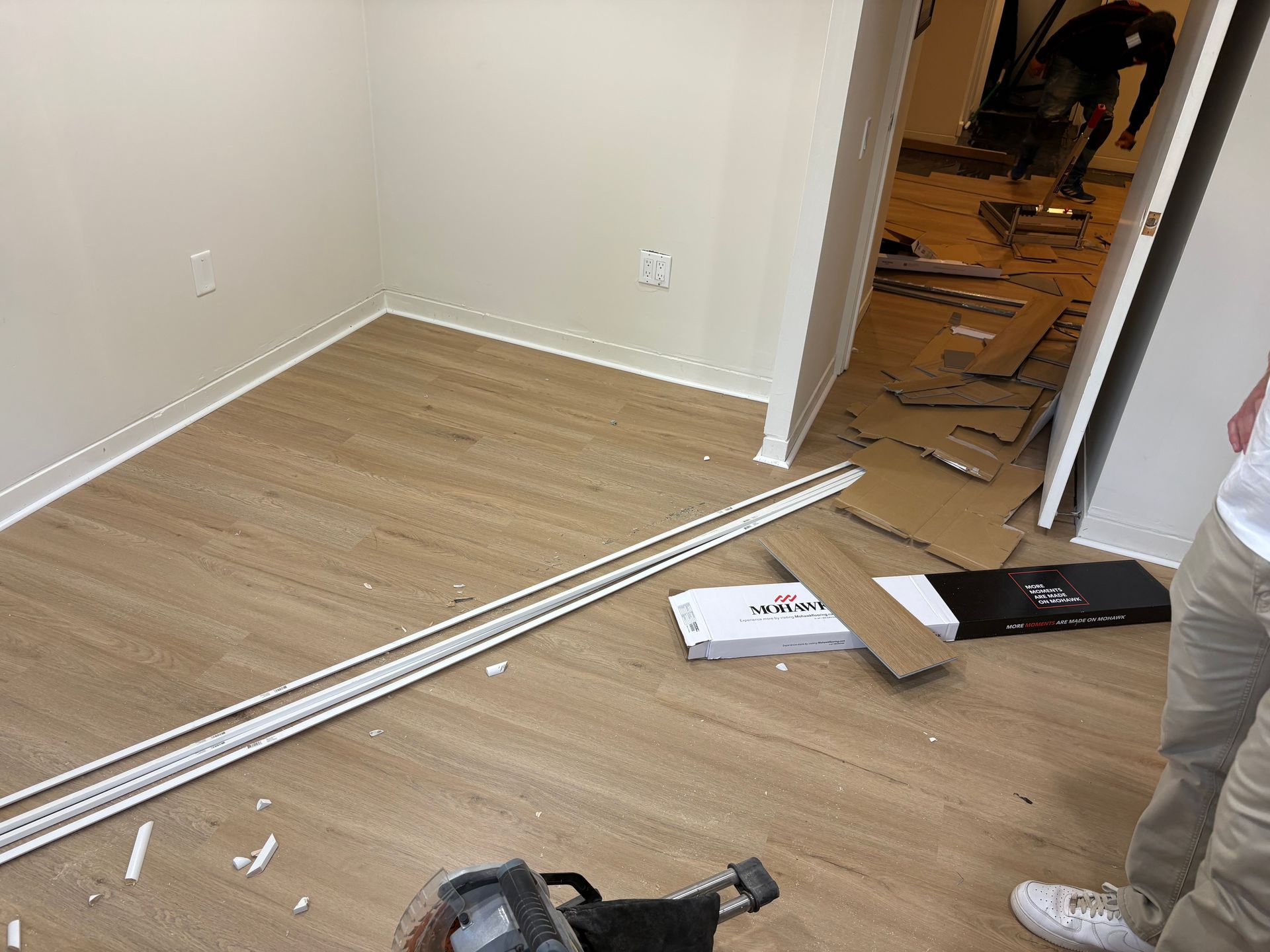 Partially renovated room with wood floor planks, trim pieces, tools, and a worker’s legs visible at right.