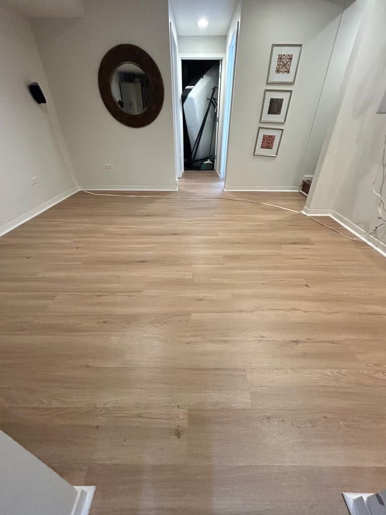 Empty hallway with light wood flooring, white walls, round mirror, and framed art near a doorway.