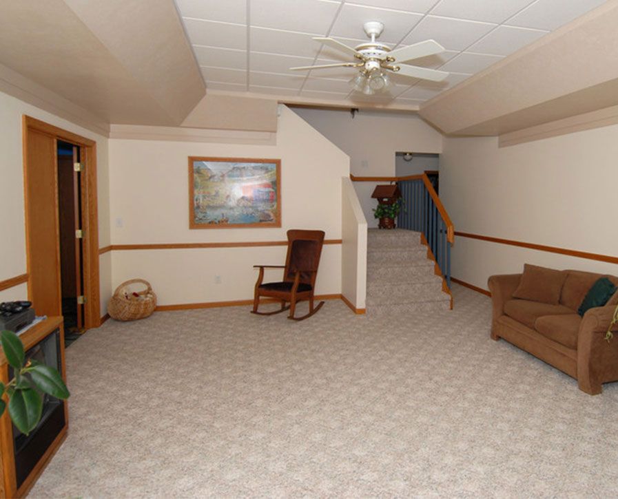Spacious beige living room with carpet, couch, rocking chair, staircase, and ceiling fan.