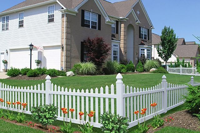White picket fence bordering a suburban house with a green lawn and bright flowers on a sunny day.