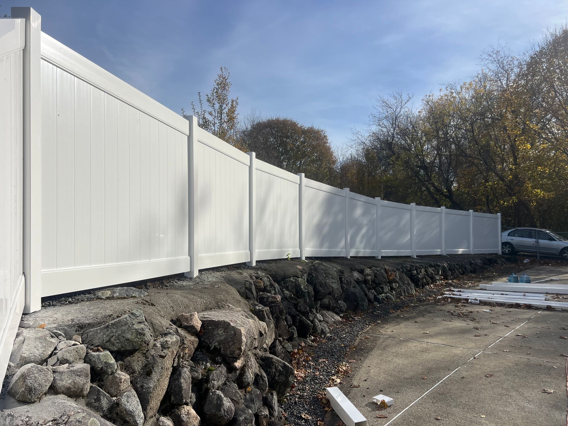 White vinyl fence along a rocky ledge, trees in the background, a sunny day.