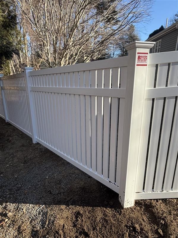 White vinyl fence along a dirt path, with trees in the background under a blue sky.