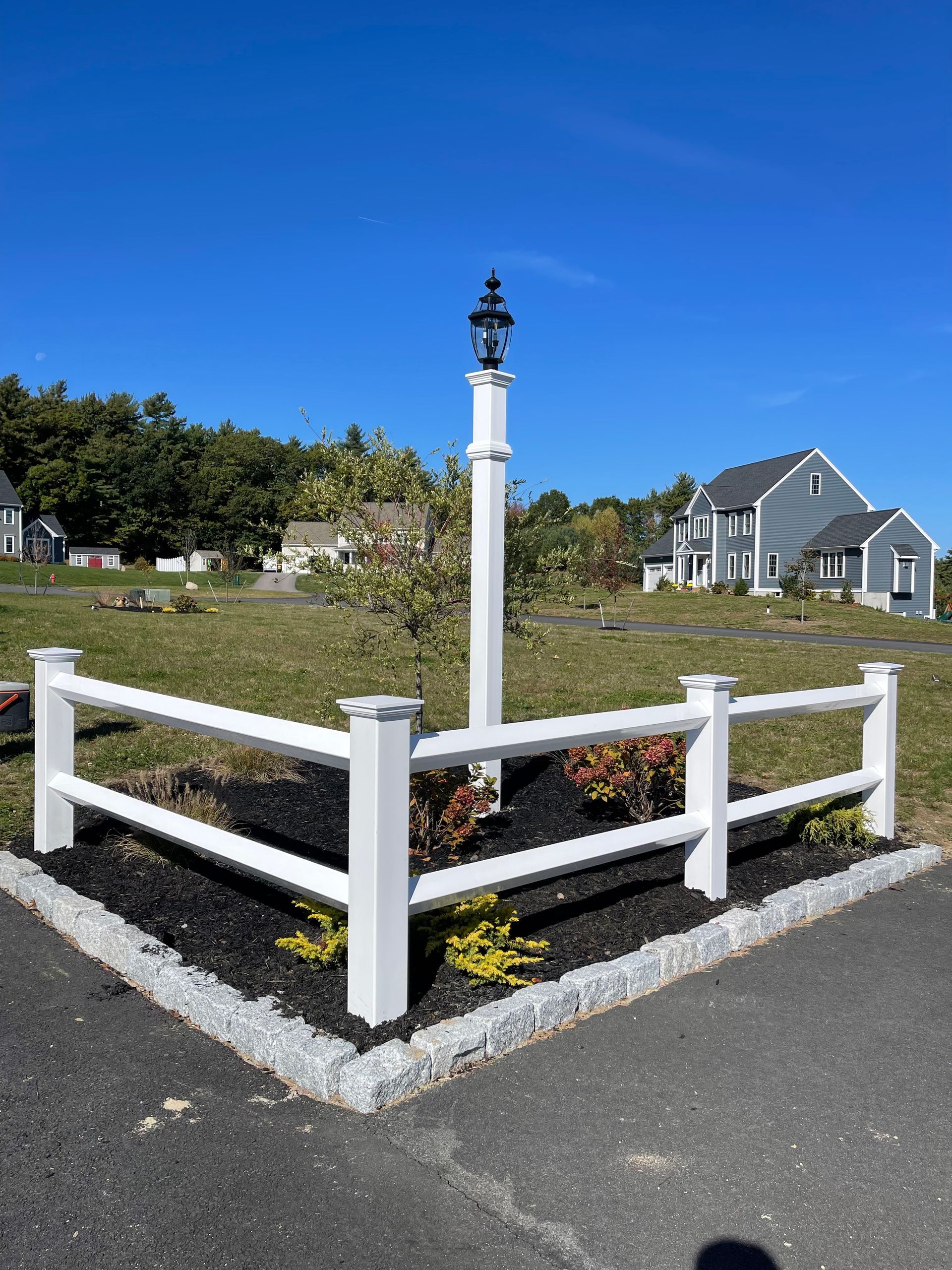 White fence and lamppost on a corner lot with a two-story gray house in the background on a sunny day.