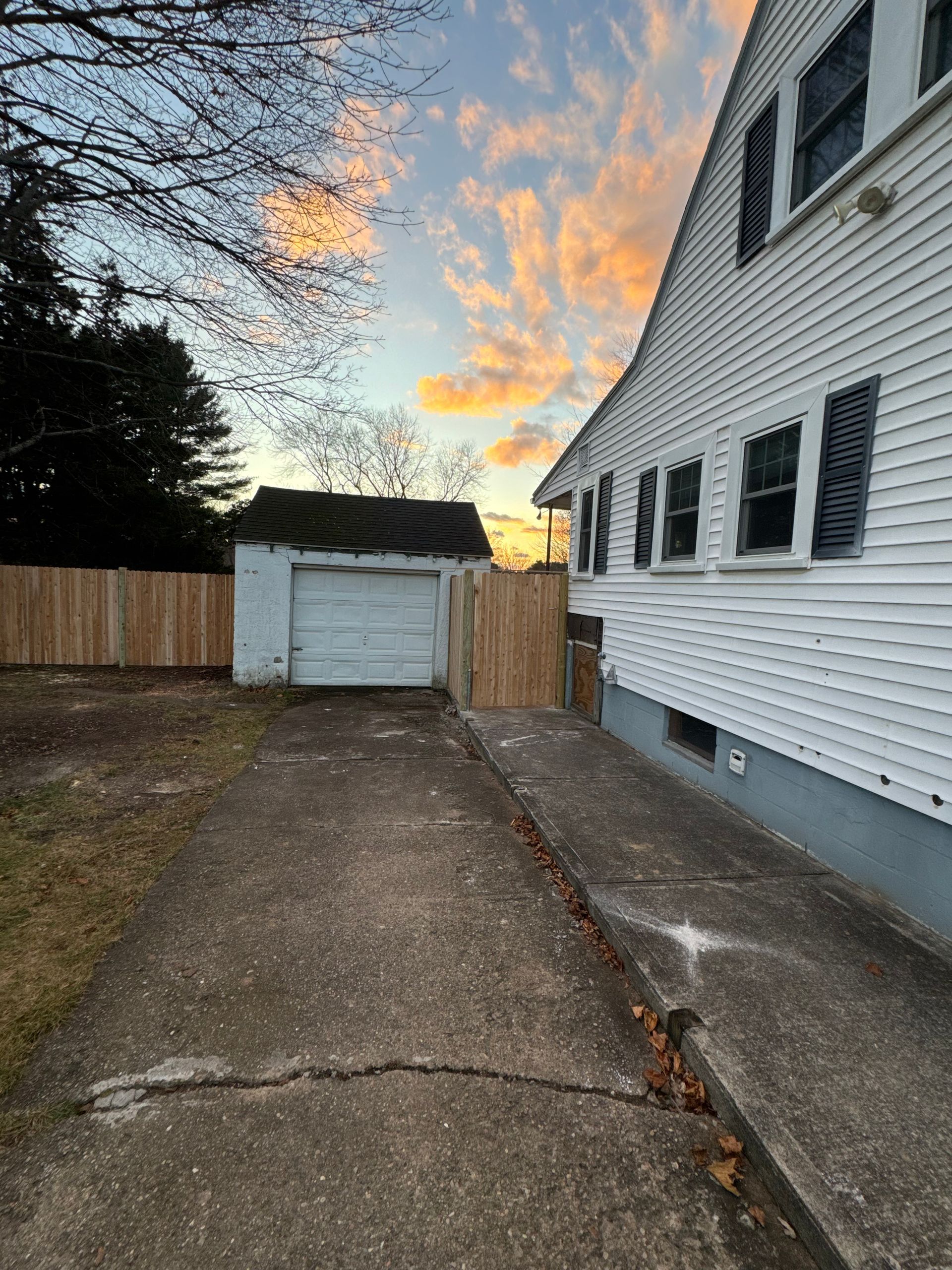 Backyard with driveway, white house, garage, and wooden fence against a sunset.