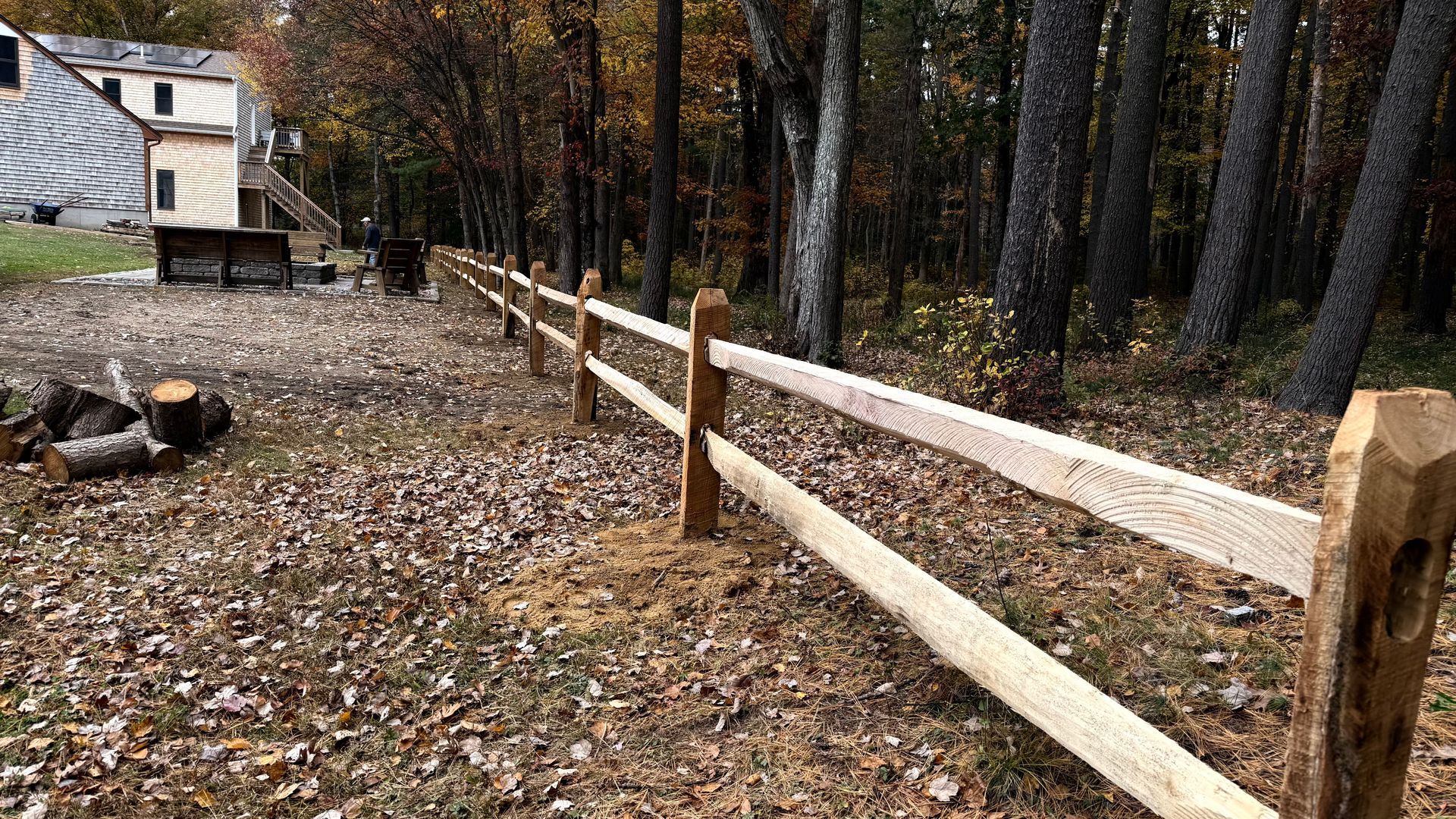 Wooden split-rail fence in a wooded area with a building in the background. Autumn leaves on the ground.
