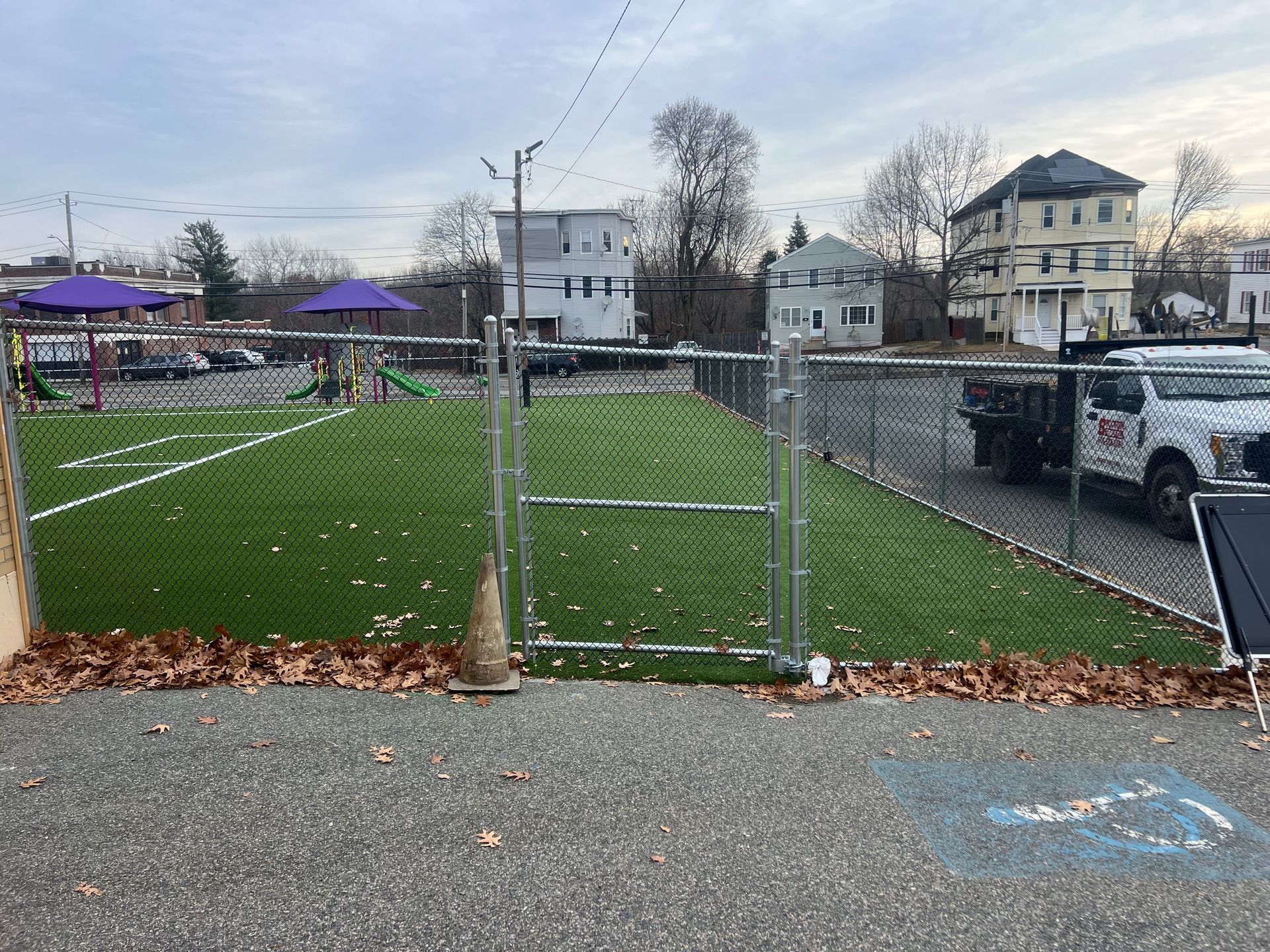 Fenced-in area with artificial turf, gate open, residential buildings in background, cloudy sky.
