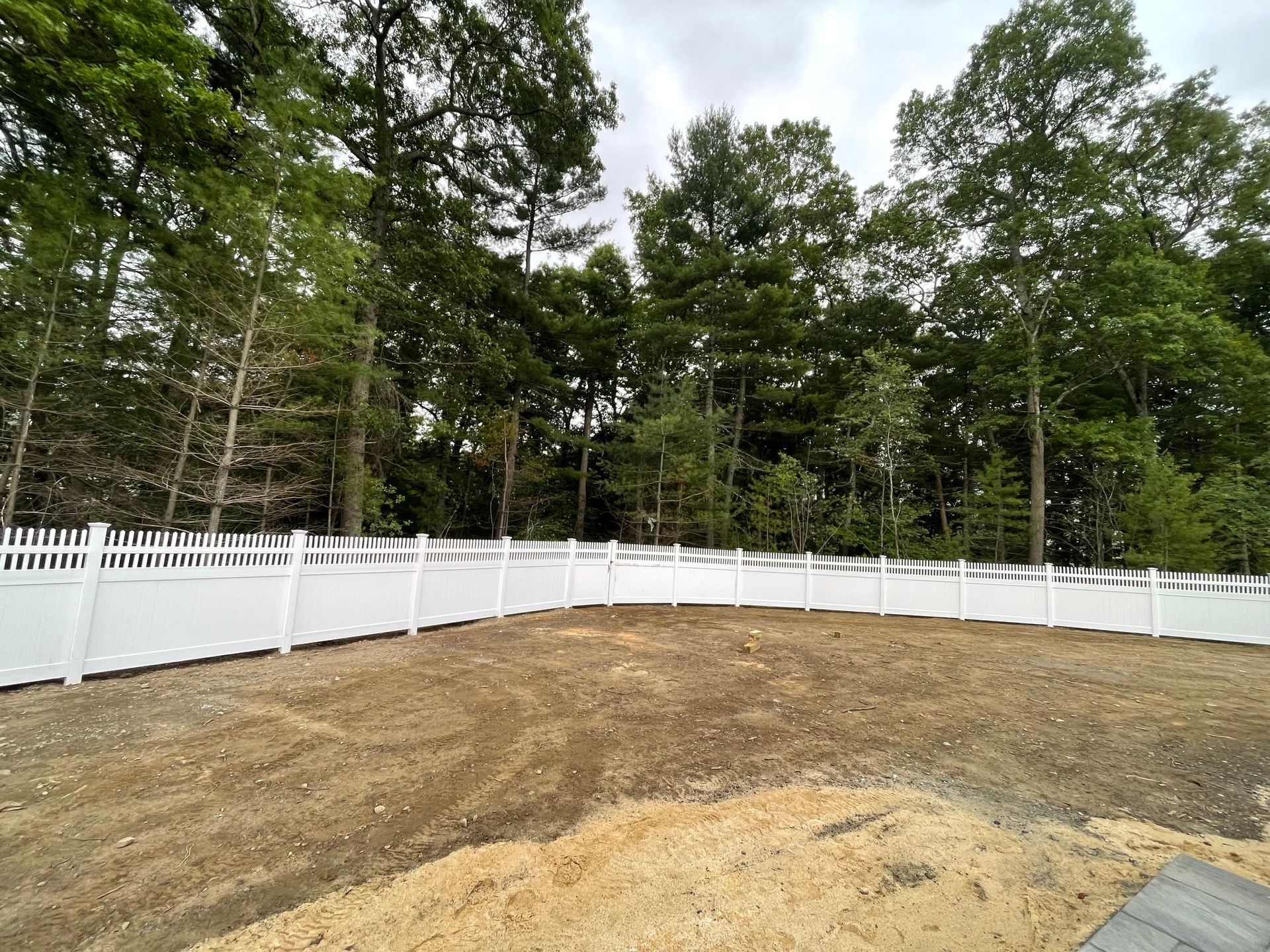 White picket fence surrounding a cleared, dirt yard with trees in the background under an overcast sky.