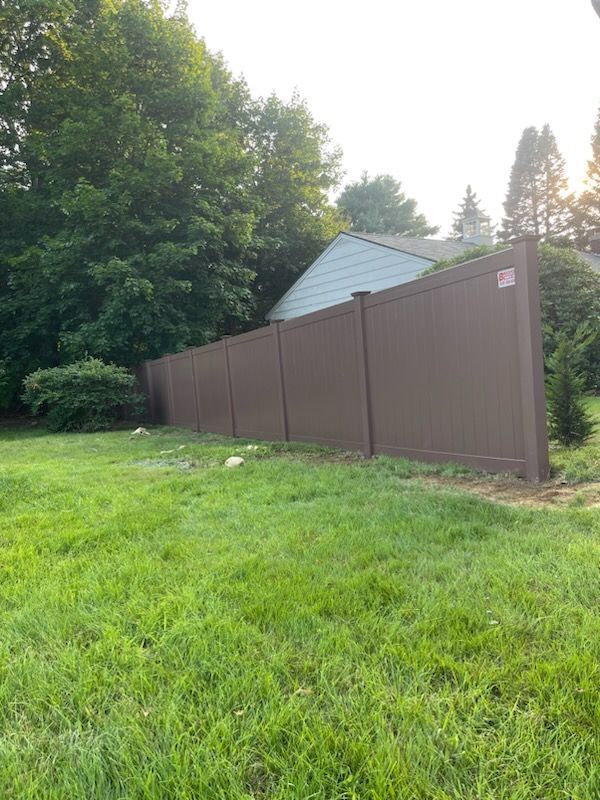 Brown vinyl fence in a grassy yard, with trees and a house in the background.