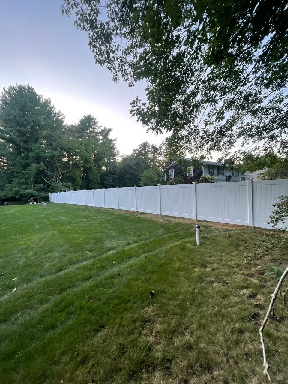 White vinyl fence along green lawn, trees in background, overcast sky.