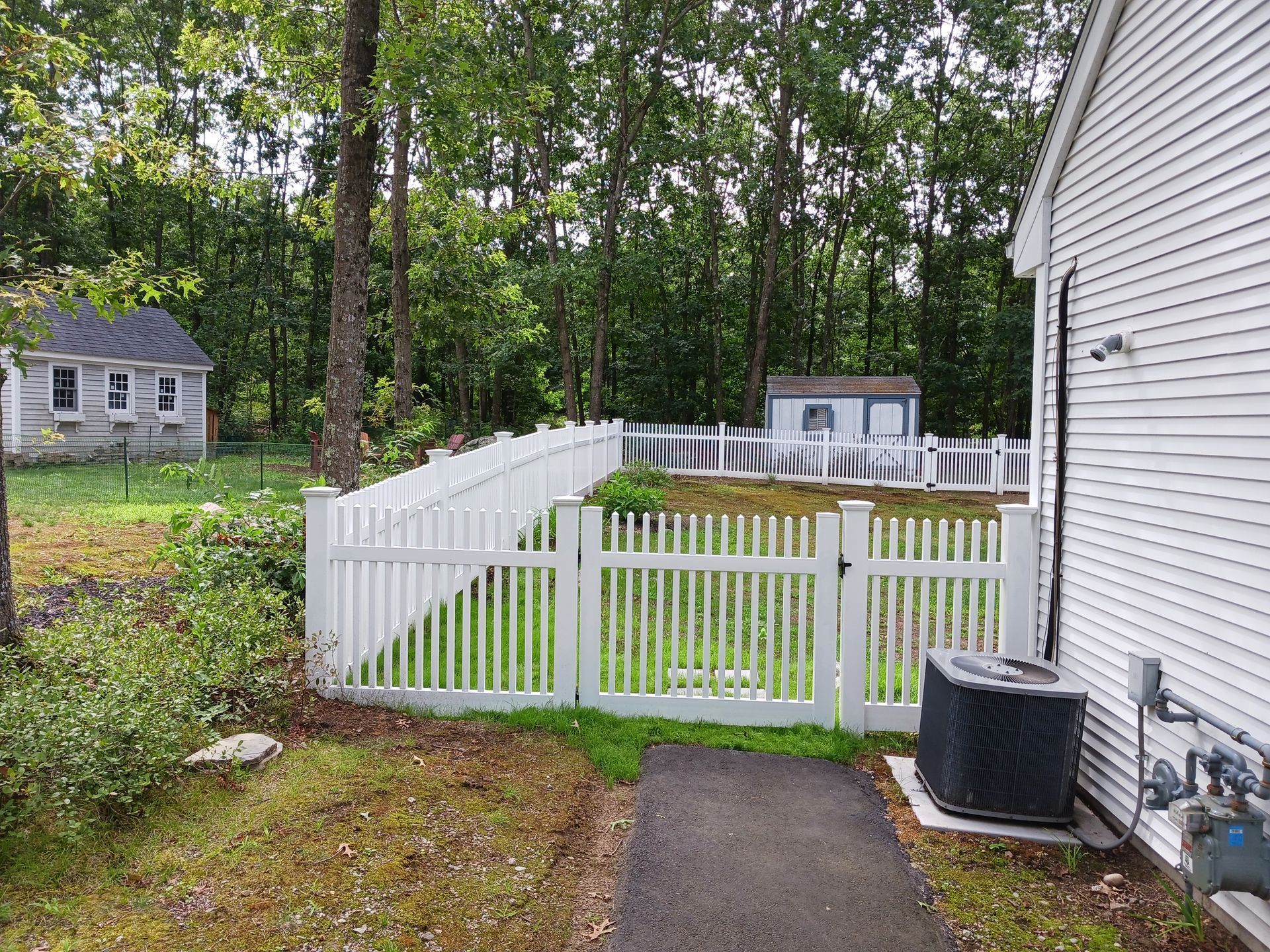 White picket fence encloses a small yard with trees, a shed, and a house. An air conditioning unit is outside.