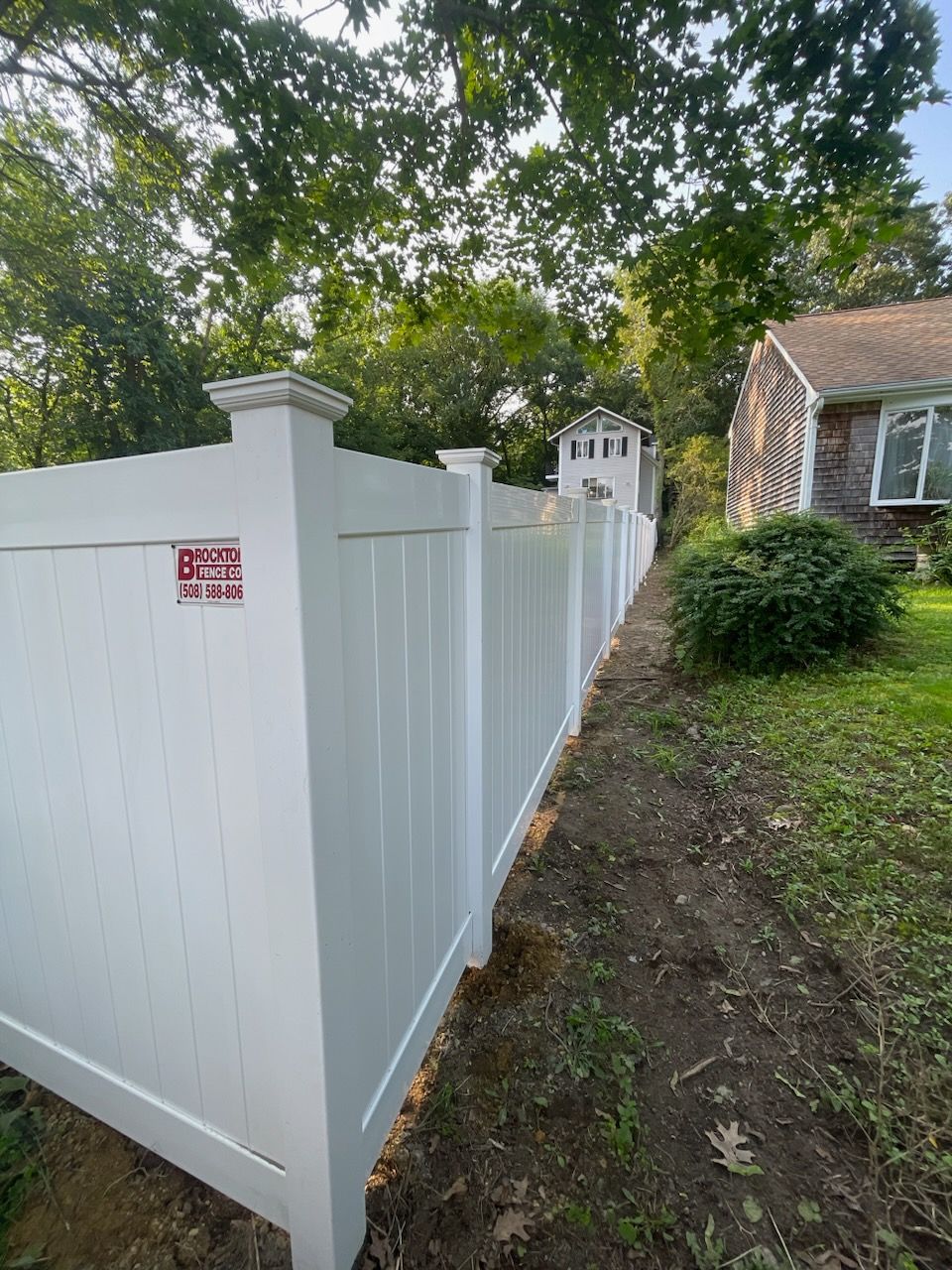 White vinyl fence along a dirt path, with a house and shed visible. Trees are in the background.