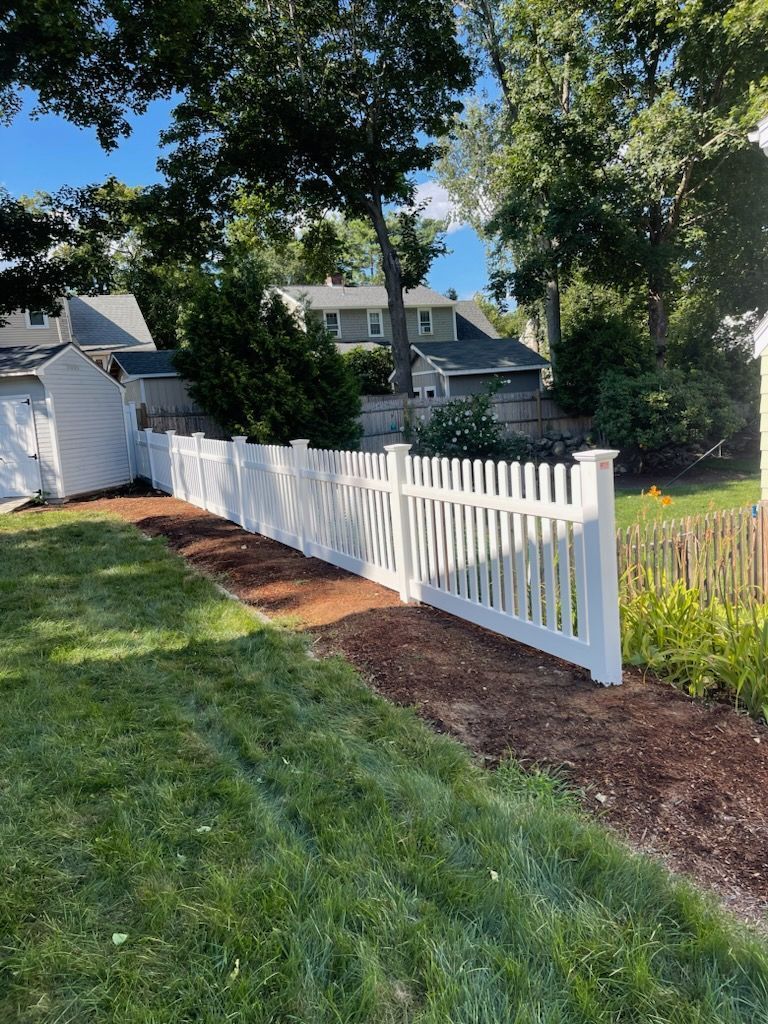 White picket fence borders a backyard with green grass and mulch. Houses and trees are in the background.