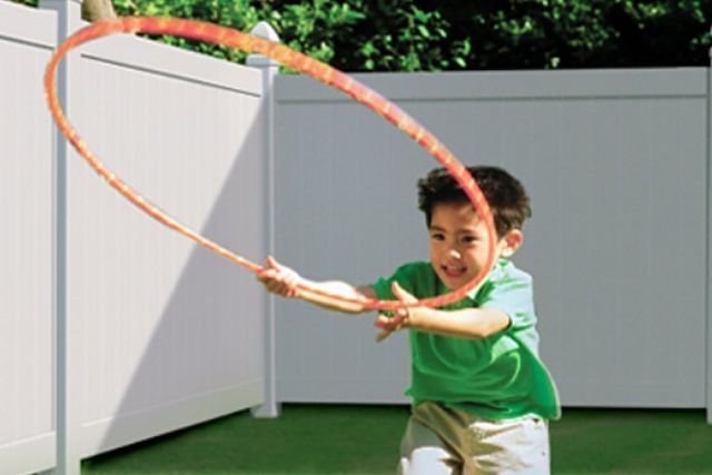 Boy in green shirt throwing a hula hoop outdoors on green turf. White fence in the background.