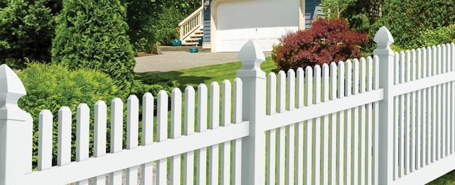 White picket fence in front of a house, green lawn, trees, and red bush.