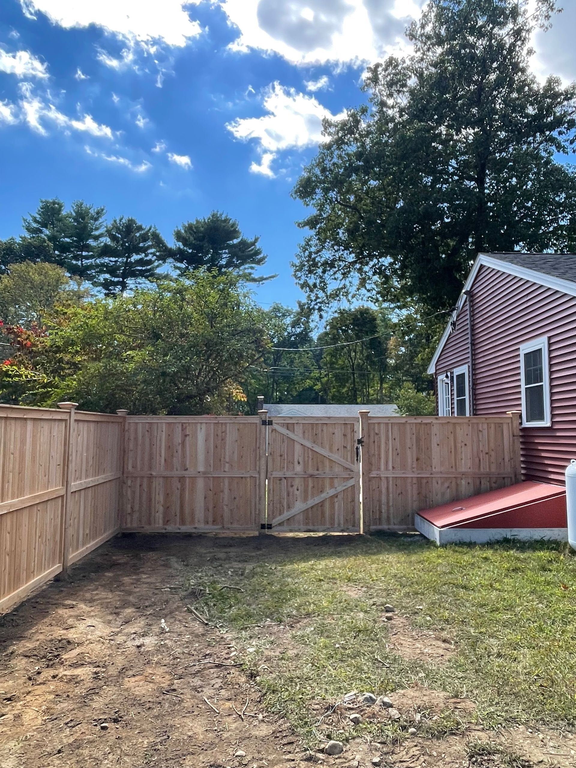 Wooden fence with gate surrounds a small backyard beside a red house; blue sky.