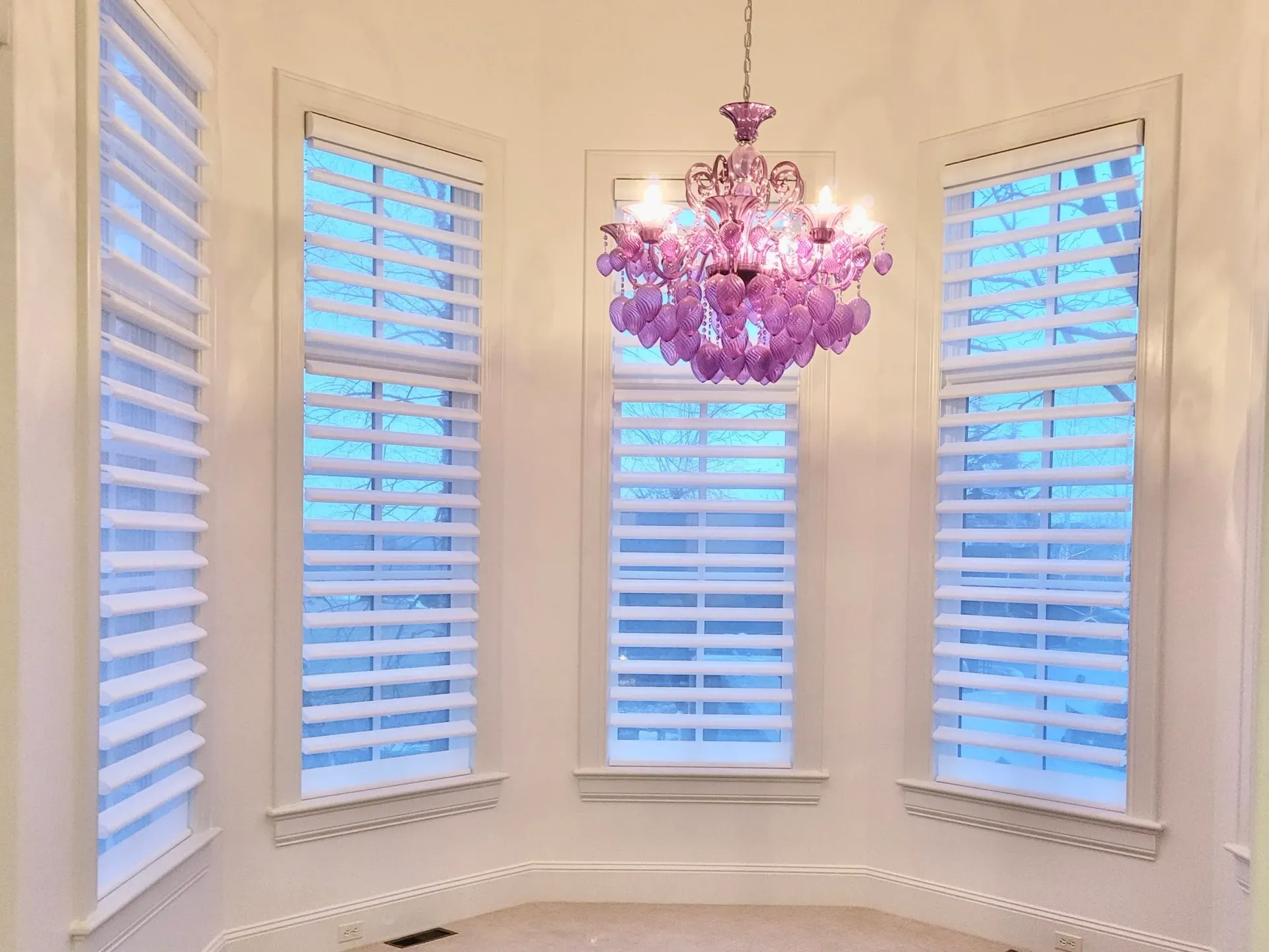 Pink chandelier hangs in a room with three shuttered windows and a curved wall.
