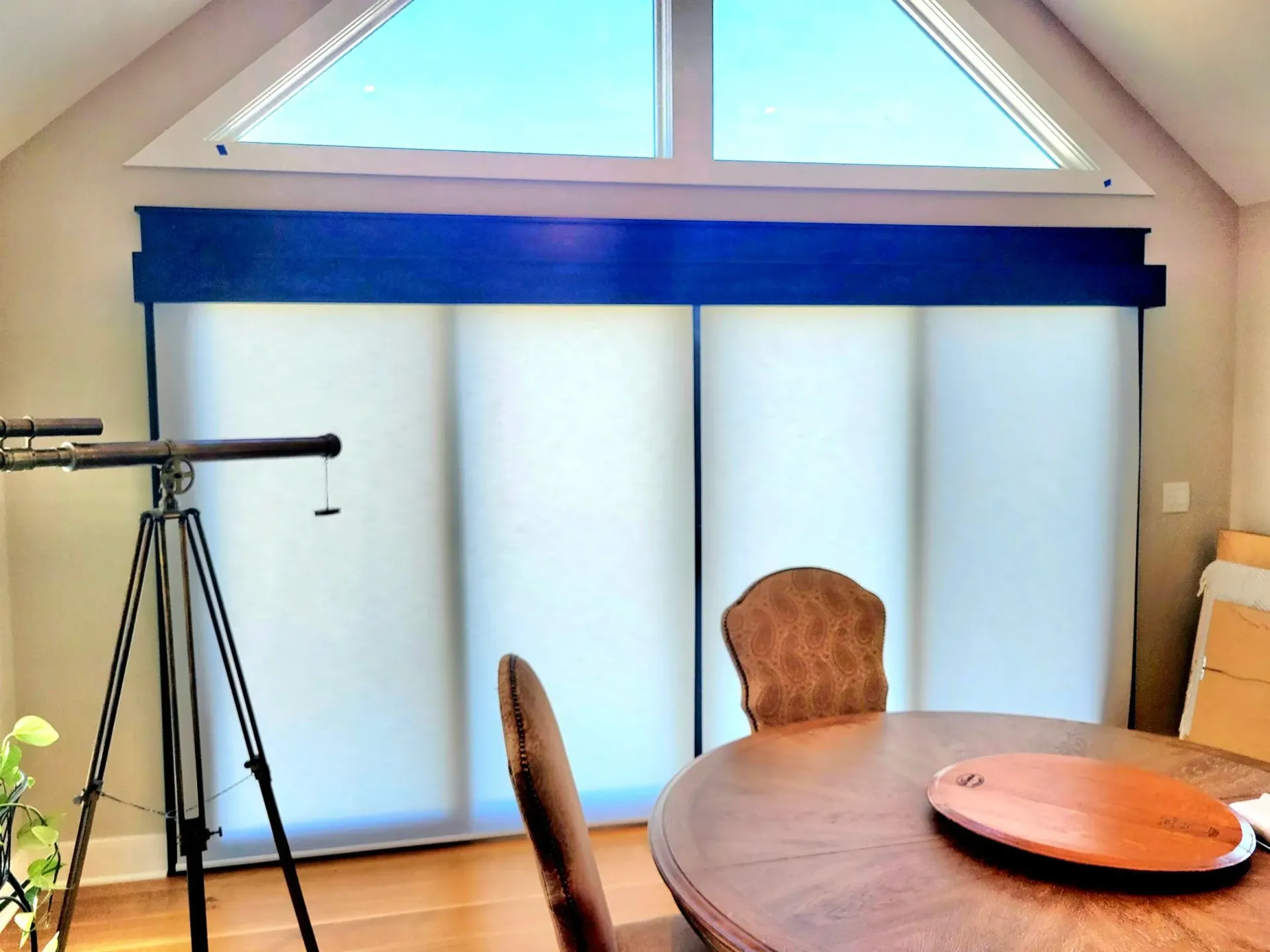 Interior with large window covered by white blinds, blue valance, and a telescope on a tripod.