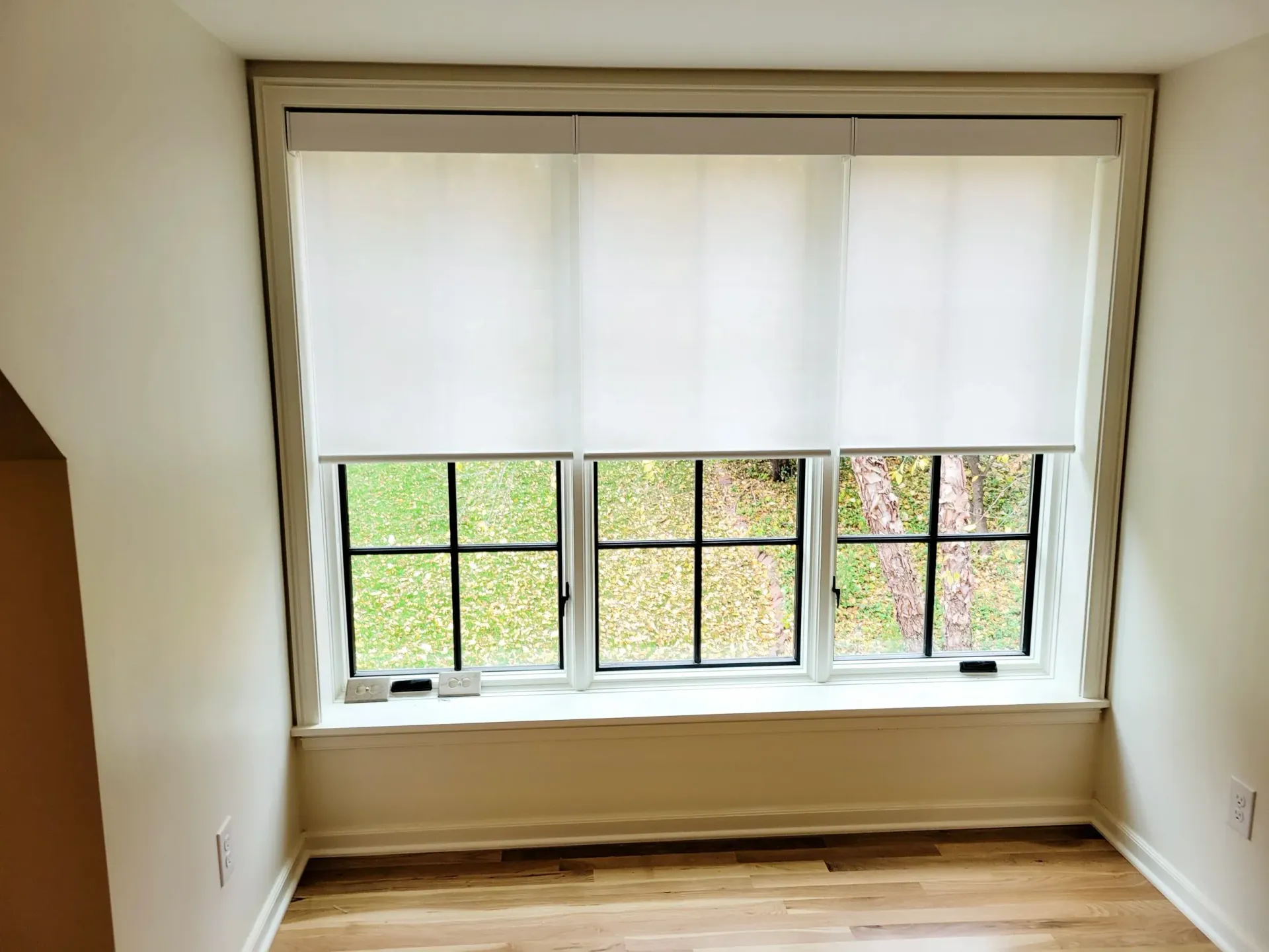 Window with three panes, each with black grid, covered by white blinds. Light wood floor and beige walls.