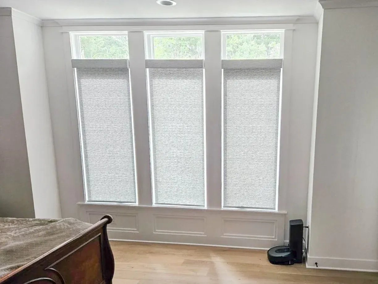Three window panels with light gray roller shades in a white room. A robot vacuum is on the floor.
