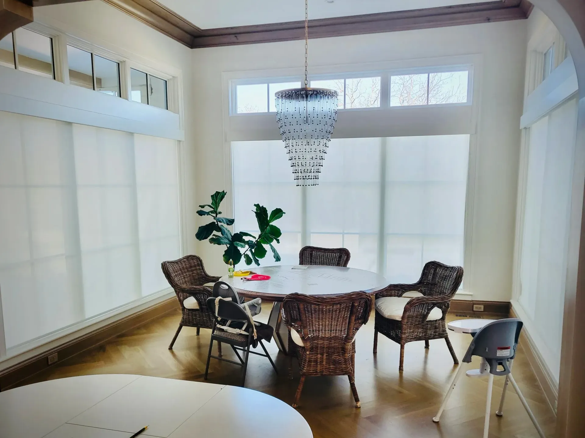 Dining room with round table, wicker chairs, chandelier, and large windows with blinds.