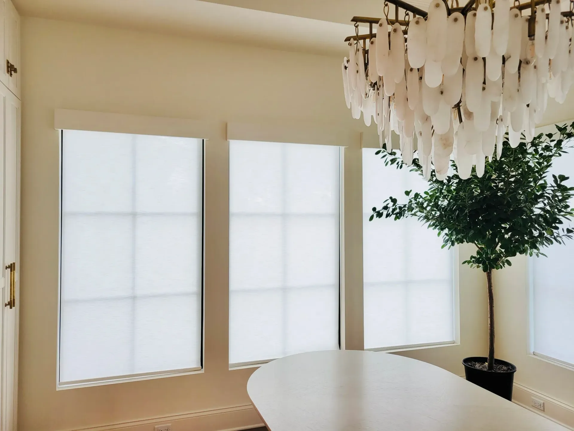 Three windows with white shades, a chandelier, and a potted tree in a light-colored room.