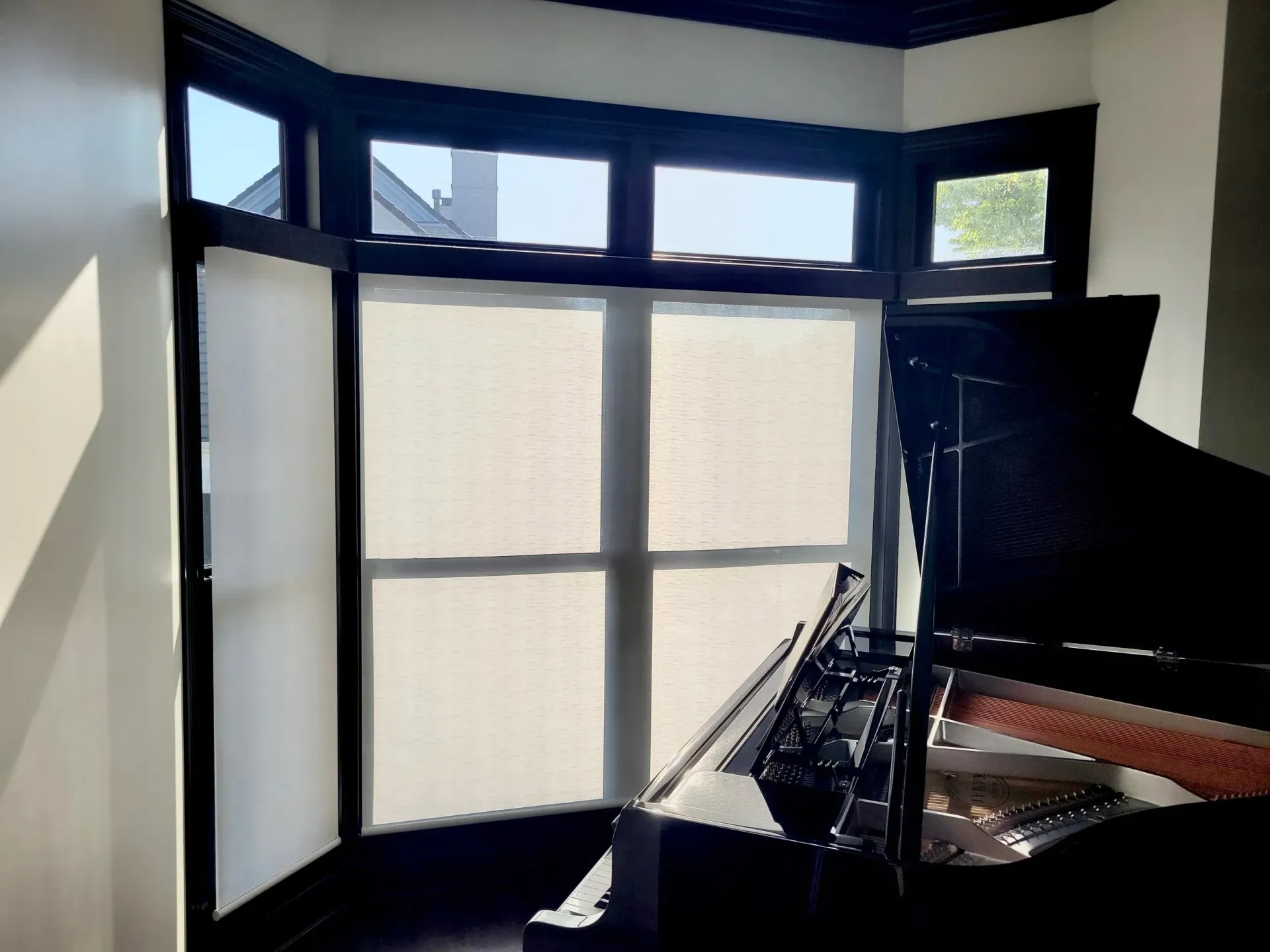 Black grand piano by a window with white shades in a room with light walls.