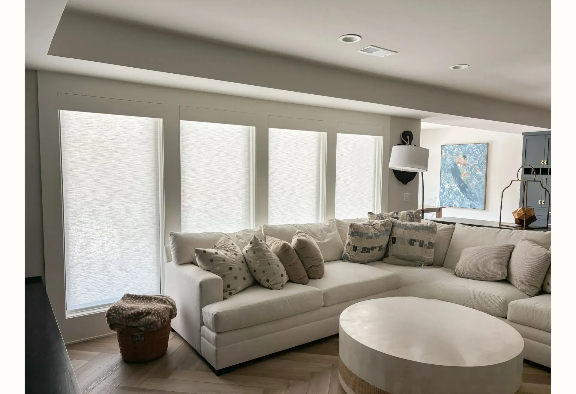 A bright living room with white sectional sofa, round coffee table, and large windows with blinds.