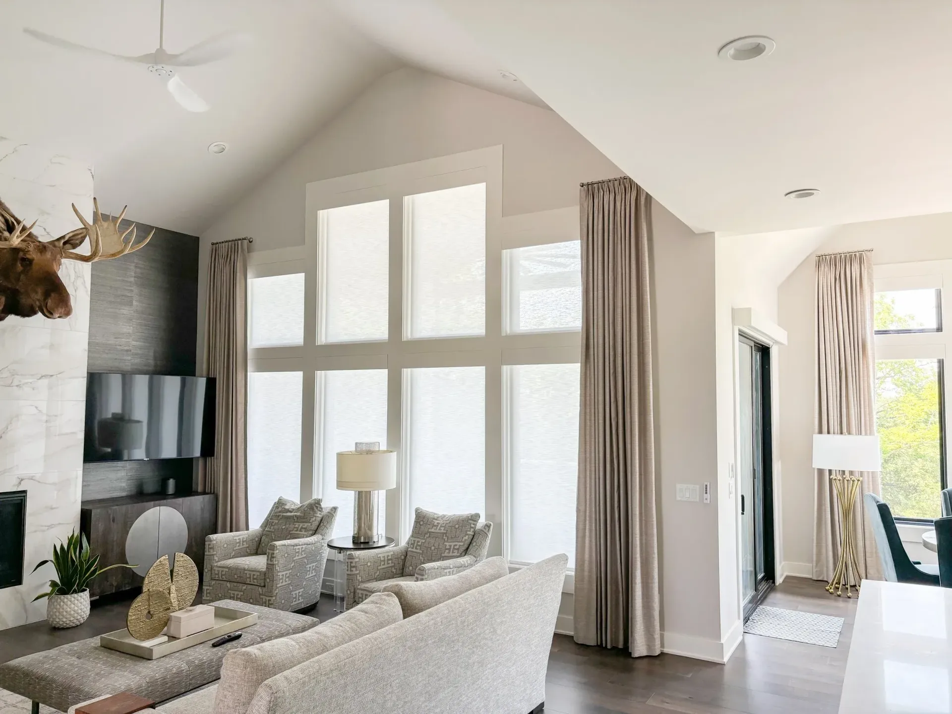 Living room with high ceilings, large windows, and neutral-toned furniture and curtains.