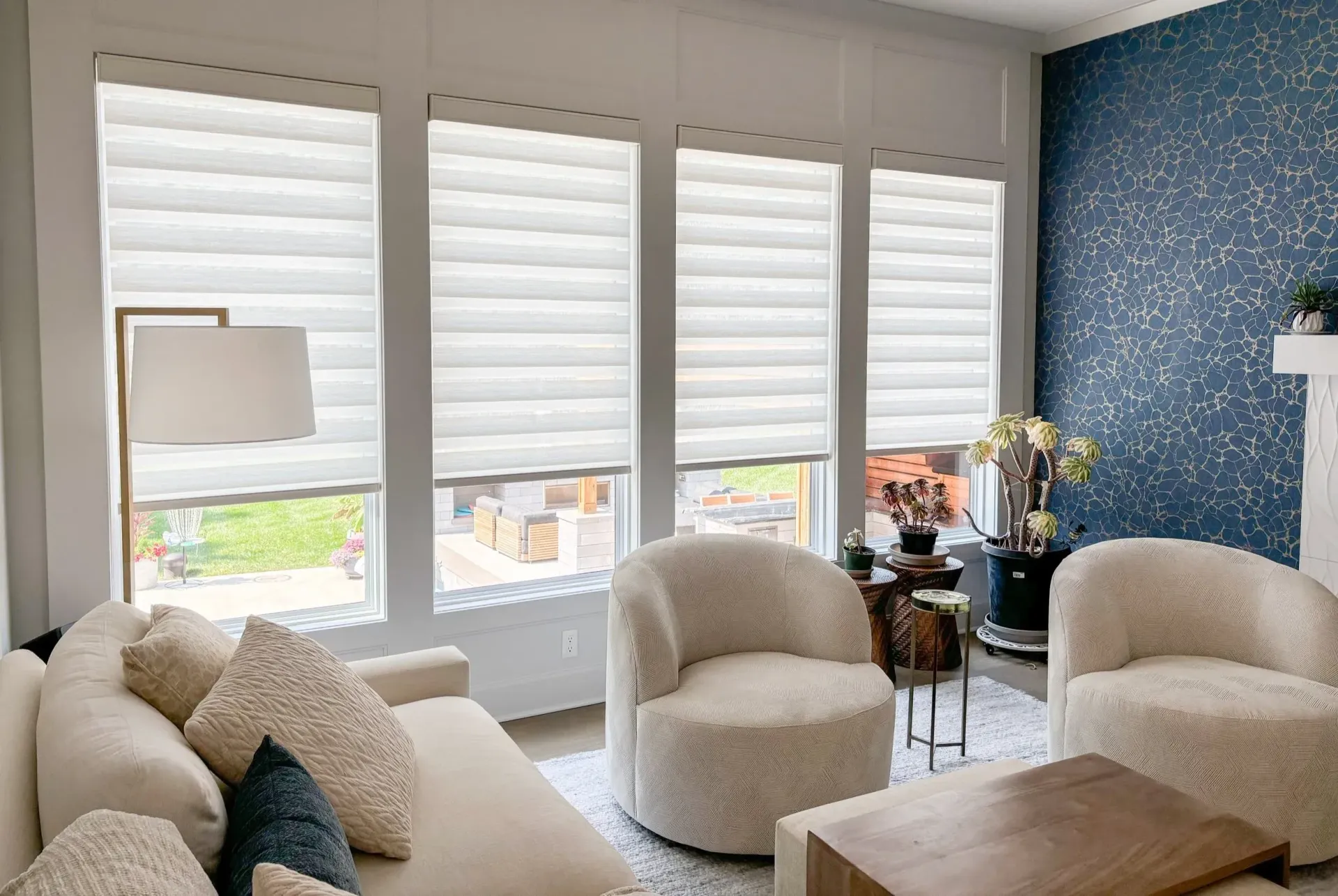 Living room with large windows, white shades, ivory sofa and chairs, blue accent wall.