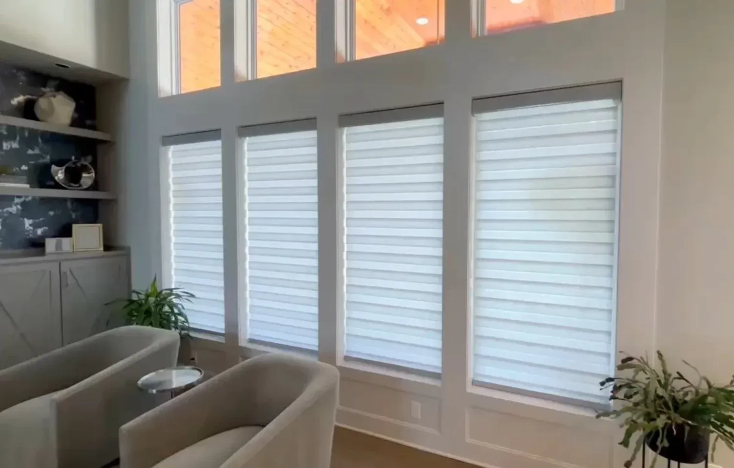 Living room with sheer white window shades, neutral armchairs, built-in shelves, and indoor plants.