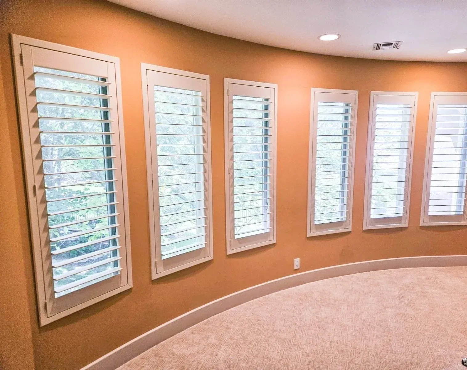 Curved room with six windows featuring white shutters and trim; peach walls, beige carpet.