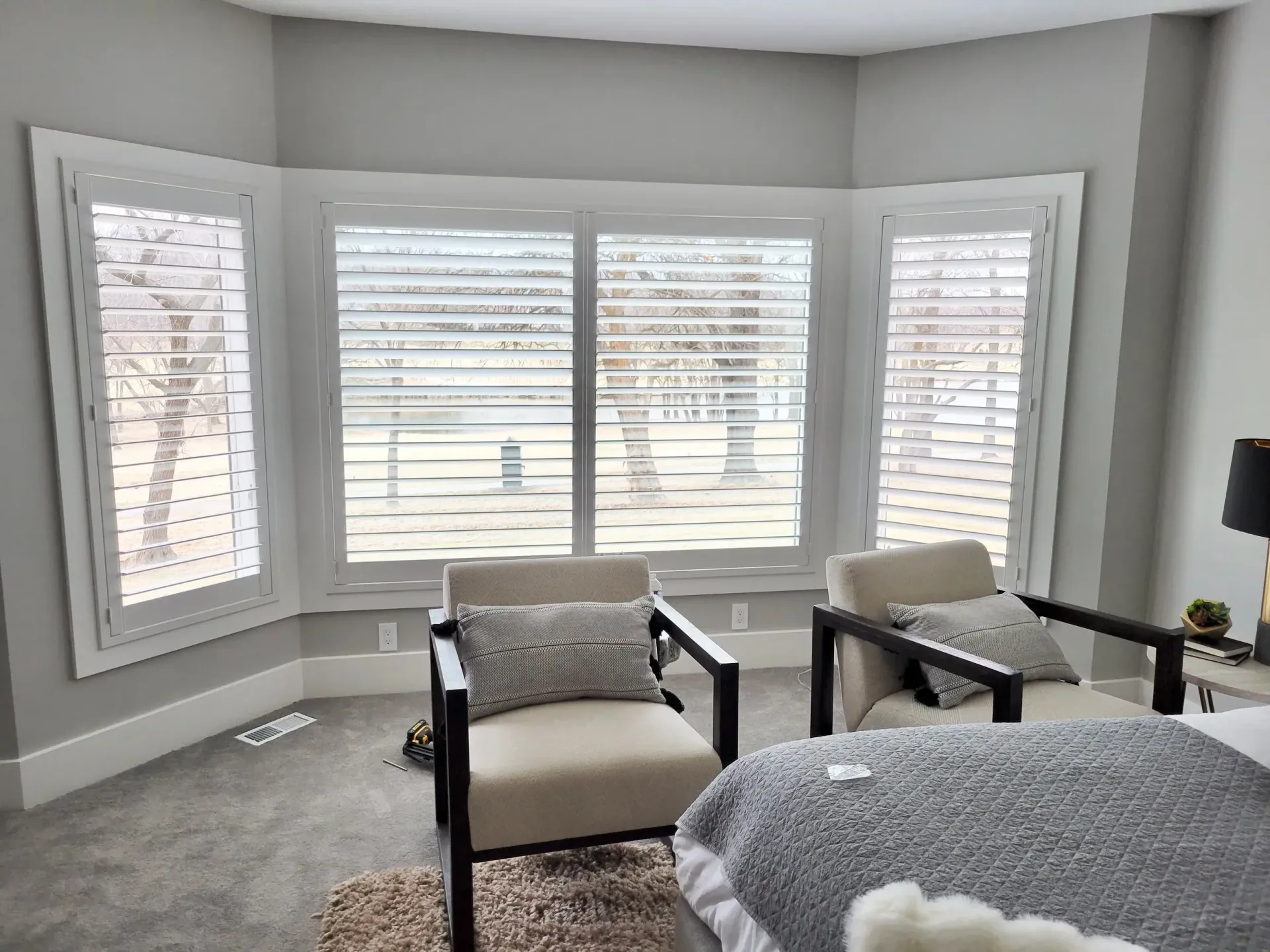 Bedroom with a bay window featuring white shutters, two armchairs, and a bed with a gray blanket.