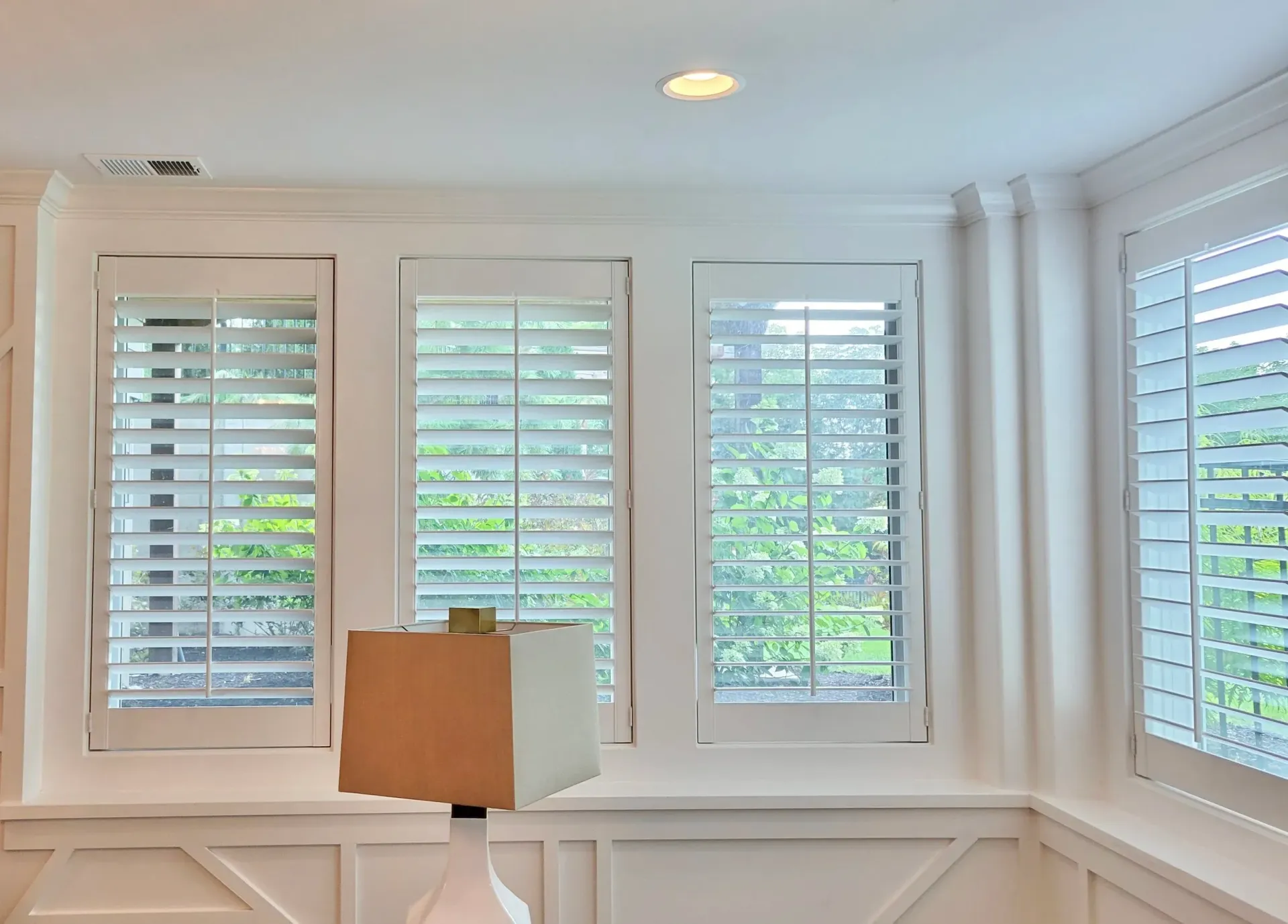 White shutters cover windows in a room with white walls and trim, a lamp sits below the windows.