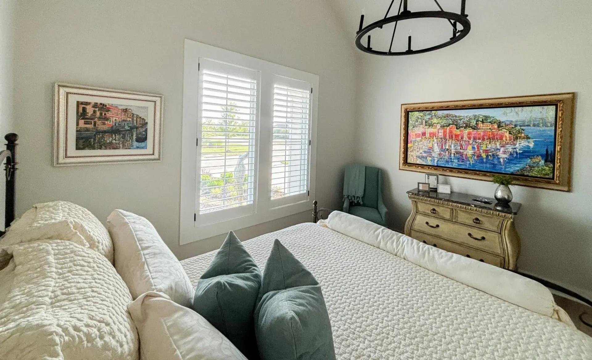 Bedroom with white shutters, bed, artwork, and a chest of drawers.