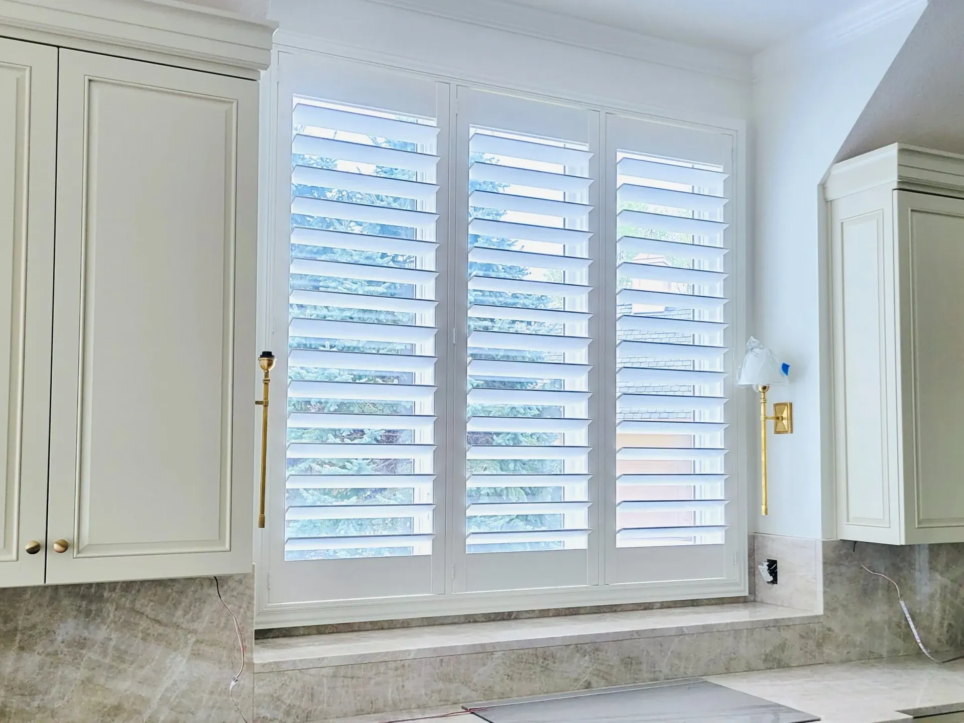 White kitchen shutters on a window between white cabinets, over a stone countertop.