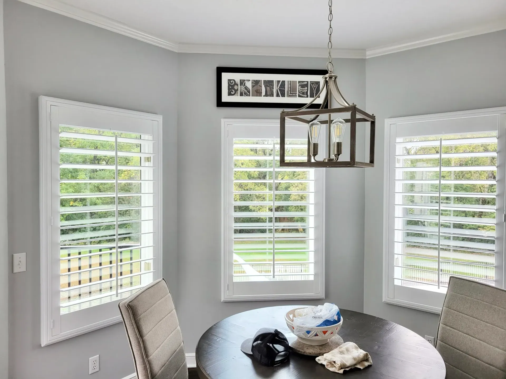 Breakfast nook with round table, three windows with shutters, and chandelier. Gray walls and white trim.